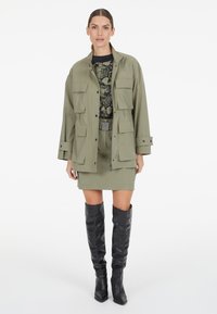 Olivgrüne Utility-Jacke mit mehreren Taschen, über einem gemusterten Oberteil und passendem olivfarbenen Rock geschichtet, ergänzt durch glänzende schwarze kniehohe Stiefel.
