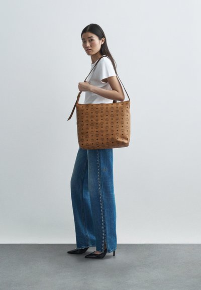 Sac à épaule beige avec un motif texturé, finition lisse et une sangle marron. Le modèle porte un t-shirt blanc et un jean large bleu avec des fentes sur les côtés.
