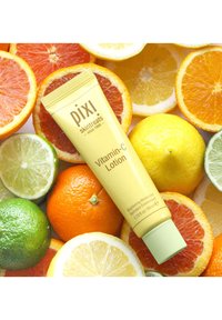Gul tube av Pixi Vitamin-C Lotion med grønn kork, omgitt av ulike sitrusfrukter, inkludert appelsiner, sitroner, lime og grapefrukt.