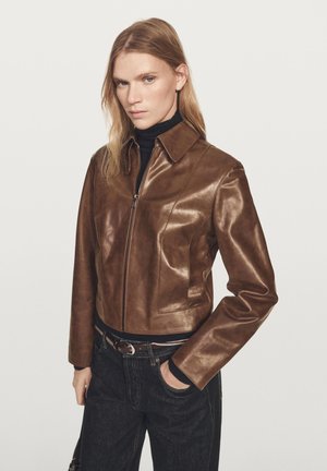 Braune Lederjacke in Kurzform mit Reißverschluss vorne, spitzem Kragen und glatter Textur. Kombiniert mit dunklen Jeans und einem schwarzen Rollkragenpullover.