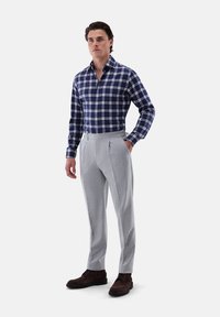 Camicia button-up a quadri navy e bianca con maniche lunghe; pantaloni grigi chiari su misura, stivali marroni, in posa eretta; sfondo bianco uniforme.