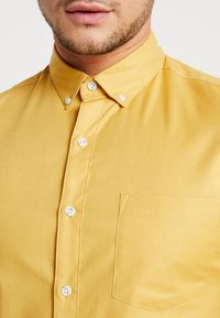 Mężczyzna w zapiętej musztardowej koszuli z kołnierzykiem typu button-down i kieszonką na piersi, ukazujący dolną część twarzy i szyję.