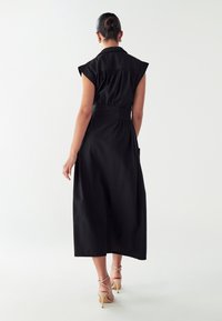 Ženska s česanimi lasmi v bunki nosi črno brezrokavno midi obleko in bež natikače z visoko peto, stoji ob preprosti beli steni.