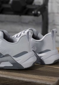 Paire de baskets athlétiques gris clair avec languettes et logos de marque sur une surface en bois, avec un arrière-plan flou.