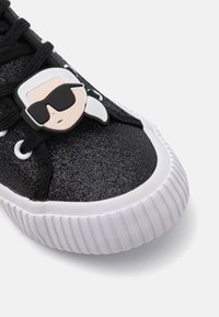 Μαύρα gliter sneakers με λευκή ελαστική σόλα, διαθέτοντας ένα μπρελόκ με καρτούν. Σχέδιο με κορδόνια, με στρογγυλή μύτη και υφή επιφάνειας.
