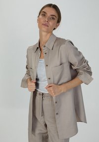 Beige Hemd mit Knöpfen aus glattem Stoff, mit einer Brusttasche, hochgekrempelten Ärmeln und einer lockeren Passform. Darunter wird ein graues Tanktop getragen.