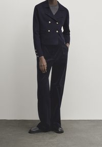 Granatowa skórzana kurtka z podwójnym zapięciem na złote guziki, zestawiona z szerokimi spodniami z aksamitu w dopasowanym kolorze, z bocznymi kieszeniami.