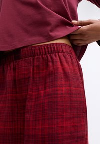 Røde ternede loungebukser med elastisk talje, lavet af blødt stof. En maroon top er delvist løftet for at afsløre buksernes talje.