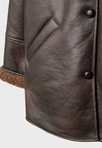 Tummanruskea nahkatakin hiha ja etupaneeli, joissa on vinottain oleva tasku, mustat pyöreät napit ja näkyvissä ruskea karvahihna.