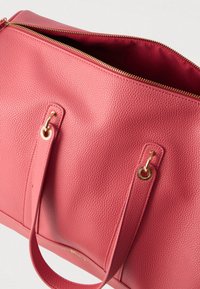 Rózsaszín texturált bőrtáska arany cipzárral, részben nyitva, két fogantyúval, amelyek arany karikákhoz csatlakoznak, és textillel bélelt belsővel.