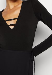 Zwarte lange mouwen bodysuit met een diepe V-halslijn en twee decoratieve banden, en een bijpassend zwart elastisch lijfje. Glad, aansluitend textuur.