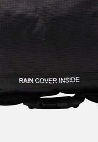 Zwarte stof met een wit label met de tekst "REGENHOES BINNENIN" boven een zwart plastic gesp of sluiting.