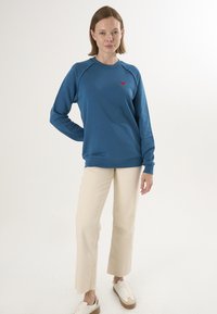 Niebieska bluza z raglanowymi rękawami, zdobiona małym czerwonym sercem wyszytym na piersi, w połączeniu z kremowymi spodniami o szerokich nogawkach i trampkami.