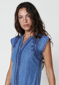 Camicia di jeans con maniche corte e arrotolate, chiusura frontale con bottoni e ricami intricati lungo il collo e le spalle in un colore azzurro chiaro.