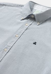 Košile na knoflíky s tenkými vertikálními šedými pruhy a malým vyšitým černým logem na hrudi, položená na bílé pozadí.