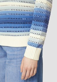 Strickpullover mit horizontalen Streifen in verschiedenen Blautönen und Lochmuster-Details. Rippbündchen in Cremefarbe. Sichtbare Jeans.