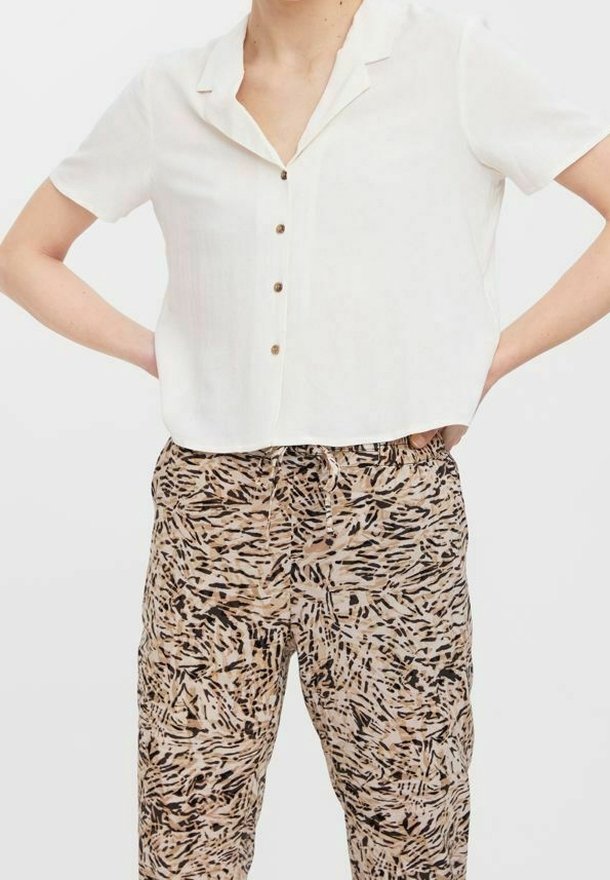Personne portant une chemise blanche à manches courtes boutonnée et un pantalon beige taille haute avec imprimé abstrait noir, les mains posées sur les hanches.