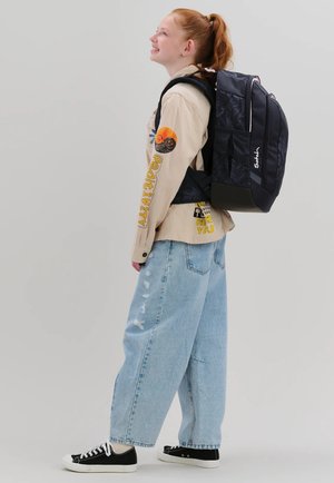 Red-haired person wearing beige shirt with "Positivity" text, light blue wide jeans, black sneakers, and carrying a large black backpack.