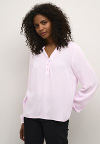 Chemise à manches longues rose clair avec un col en V et trois boutons, en tissu texturé, associée à un pantalon noir.