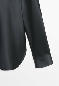 Chemise noire à manches longues avec un ourlet arrondi et un large poignet, fabriquée dans un tissu lisse et légèrement brillant.