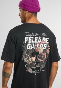 Černé bavlněné tričko s barevným grafickým potiskem kohouta na zadní straně, s nápisy "Tradición Viva" a "Pelea de Gallos" v bílé barvě.