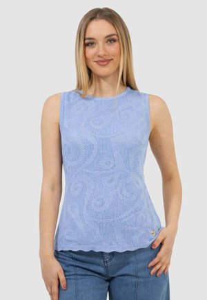 Donna con capelli biondi lisci che indossa un top a maglia senza maniche azzurro chiaro con motivo e jeans blu, sorridente con la mano sul fianco.