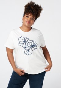 T-shirt en coton blanc avec un design floral dans les contours bleu marine. Manches courtes et encolure ronde. Porté avec un jean bleu foncé.