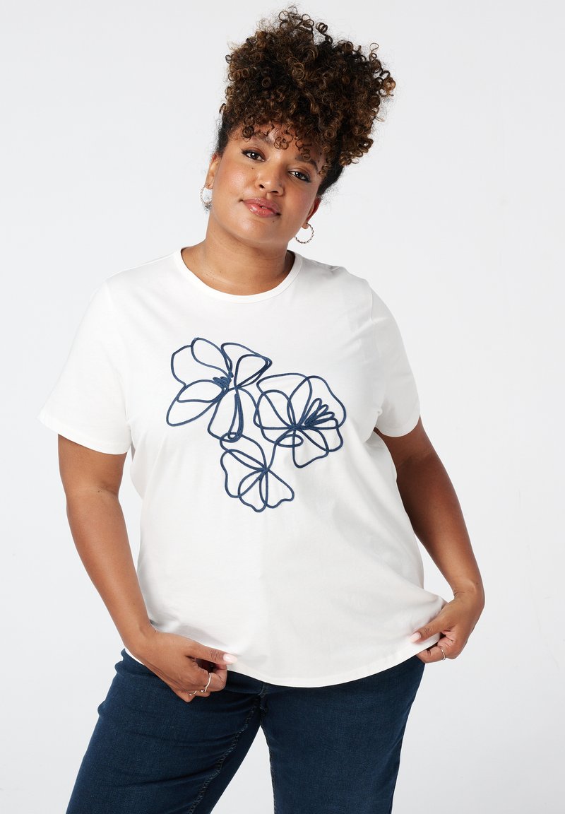 T-shirt en coton blanc avec un design floral dans les contours bleu marine. Manches courtes et encolure ronde. Porté avec un jean bleu foncé.