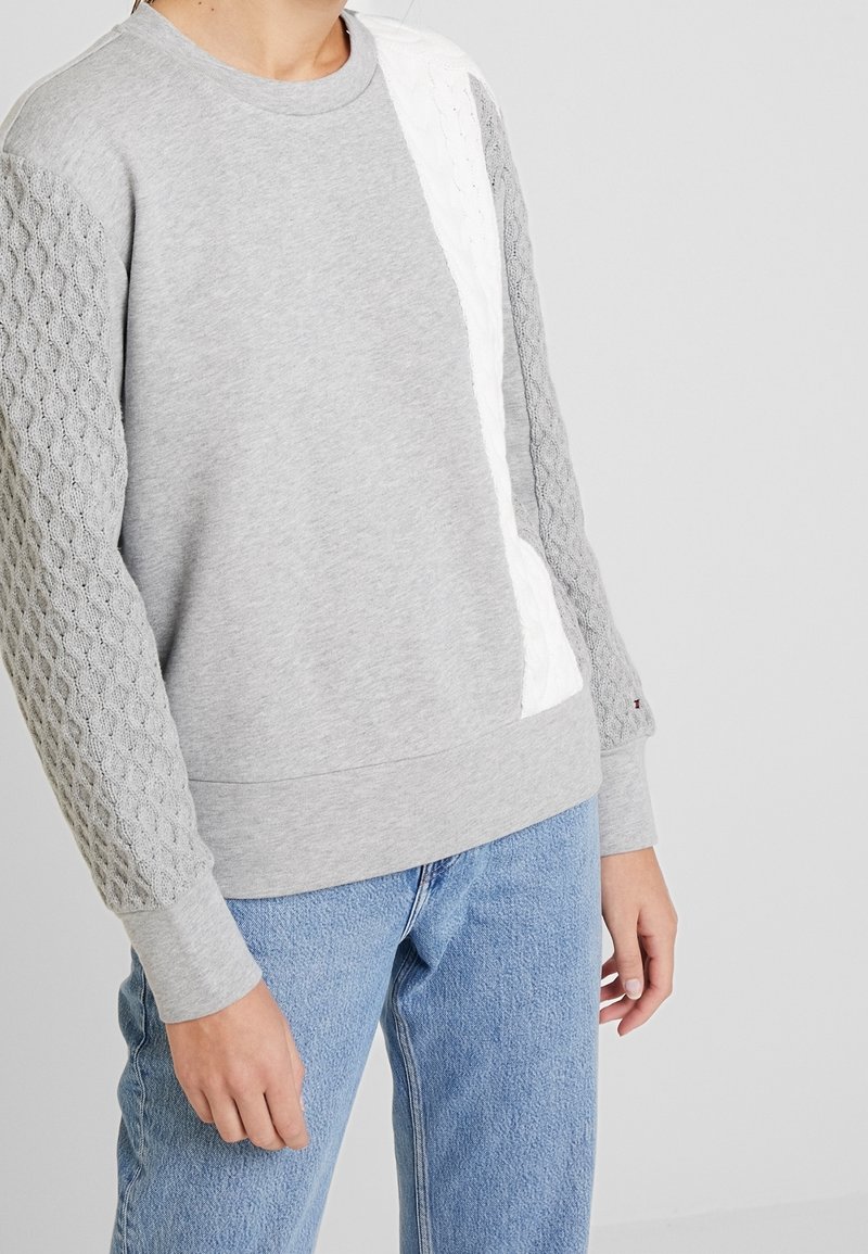 Personne portant un pull gris avec des manches en maille torsadée texturée et une rayure verticale blanche, associé à un jean bleu clair.