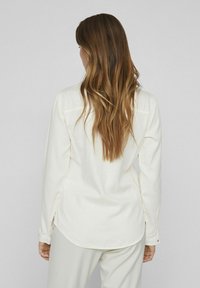 Camicia a maniche lunghe color crema chiaro realizzata in tessuto morbido, con orlo curvo, fessure laterali e polsini con bottoni e dettagli di cucitura discreti.