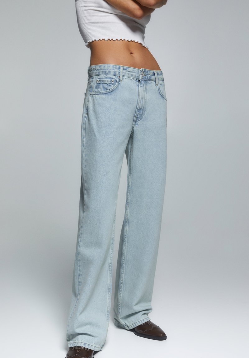 Los jeans de pierna ancha en azul claro cuentan con una cintura alta, un diseño de cinco bolsillos y sutiles detalles desgastados. La tela parece suave con una ligera textura.