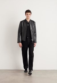 Black leather jacket with a smooth texture, side pockets, and a zip closure. Worn over a black shirt and paired with black trousers and shoes.