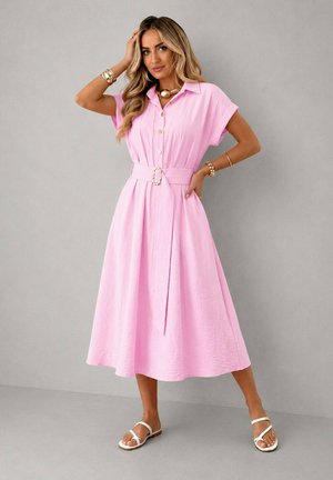 Femme portant une robe midi rose ceinturée à manches courtes, bijoux en or et sandales blanches sur un fond gris uni.