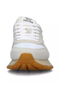 Zapatilla con parte superior de malla blanca y ante beige, suela de goma naranja y texto "LAS CHICAS CORREN MÁS RÁPIDO" en la lengüeta. Presenta cordones planos blancos.