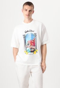 Camiseta blanca de gran tamaño con un gráfico colorido de un pueblo costero, detallada con acentos amarillos de limón y un logo vintage.