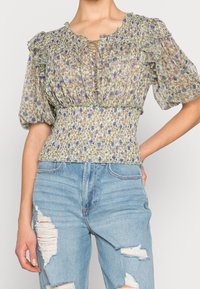 Blus med puffärmar och blommönster, med smockad midja, snörning i halsringningen och strukturerad, lättvikts tyg i gröna och lila nyanser.