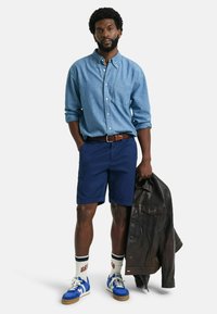 Blauwe chambray overhemd met knopen, marineblauwe shorts, blauwe sneakers met witte veters, witte sokken met rode accenten en een bruine leren jas.