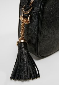 Roh černé texturované kožené kabelky se zlatým zipem, zlatou řetězovou střapcem s koženými prameny a malým gravírovaným zlatým kroužkem.