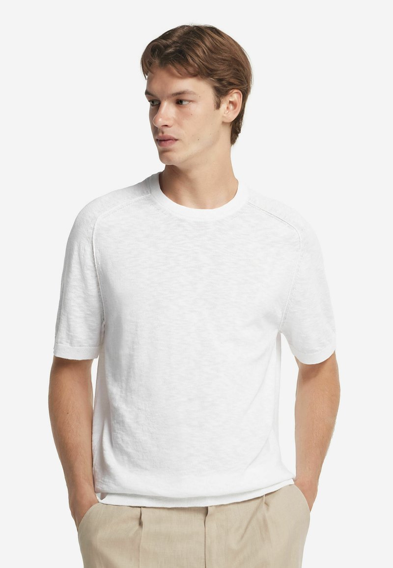 Hvid kortærmet t-shirt med rund halsudskæring, tekstureret stof og subtile stikninger på raglanærmer. Bæret med beige bukser.