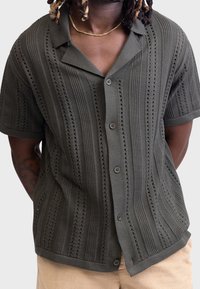 Kurzärmeliges, olivgrünes Strickshirt mit einem strukturierten Muster aus abwechselnden Streifen und Lochmustern, mit durchgehender Knopfleiste und weichem Kragen.