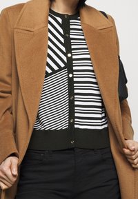 Bruine wollen jas over een zwart-wit gestreepte cardigan met knoopdetails. Zwarte broek maakt de outfit compleet. Getextureerde en gelaagde uitstraling.