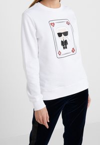 Sudadera blanca con un gráfico de una carta que muestra a un personaje con traje negro y gafas de sol; puños y dobladillo acanalados con un corte relajado.