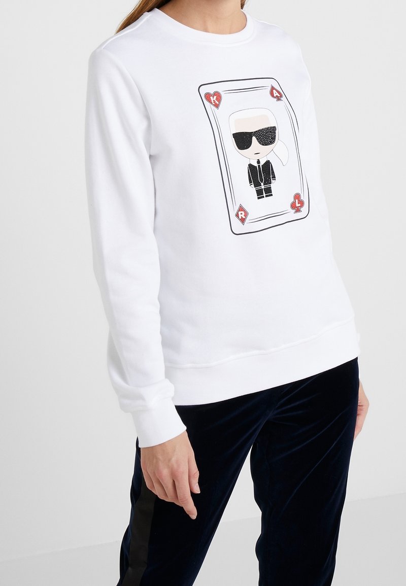 Sudadera blanca con un gráfico de una carta que muestra a un personaje con traje negro y gafas de sol; puños y dobladillo acanalados con un corte relajado.