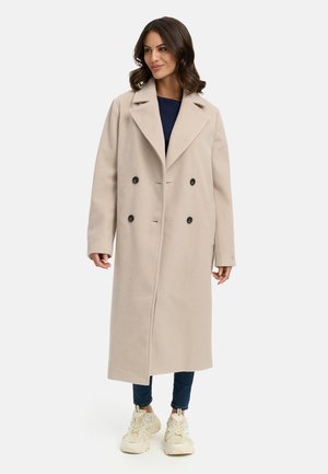 Manteau en mélange de laine beige avec un design croisé, grands revers, deux poches latérales et boutons noirs, associé à un jean bleu et des baskets blanches.