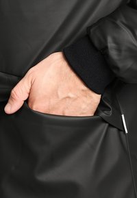 En vuxenhand delvis inskjuten i fickan på en svart jacka med ribbad mudd.