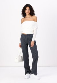 Top blanco sin hombros, de mangas largas, combinada con pantalones oscuros a rayas y zapatillas blancas. Se sostiene un pequeño bolso blanco.