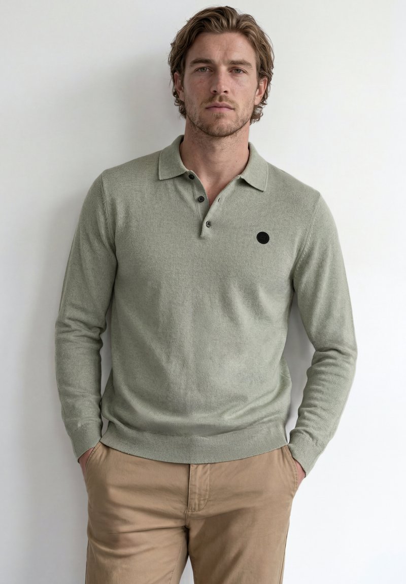 Homme aux cheveux châtain clair portant un polo manches longues vert clair et un pantalon beige, debout contre un mur blanc les mains dans les poches.