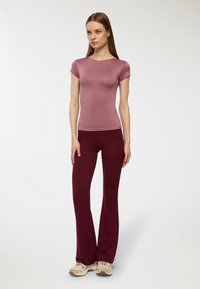 Jasnofioletowa, dopasowana bluzka z krótkim rękawem, połączona z bordo rozszerzanymi spodniami, wykonanymi z miękkiego materiału. Modelka ma na sobie beżowe buty sportowe.