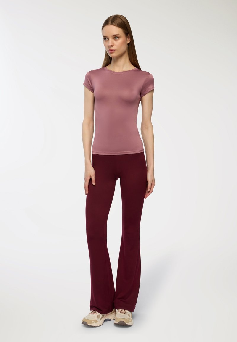 Jasnofioletowa, dopasowana bluzka z krótkim rękawem, połączona z bordo rozszerzanymi spodniami, wykonanymi z miękkiego materiału. Modelka ma na sobie beżowe buty sportowe.