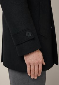 Czarny wełniany płaszcz z guzikiem przy mankietach, teksturowany materiał i dopasowany krój; ręka umieszczona blisko dołu, podkreślająca eleganckie detale projektu.